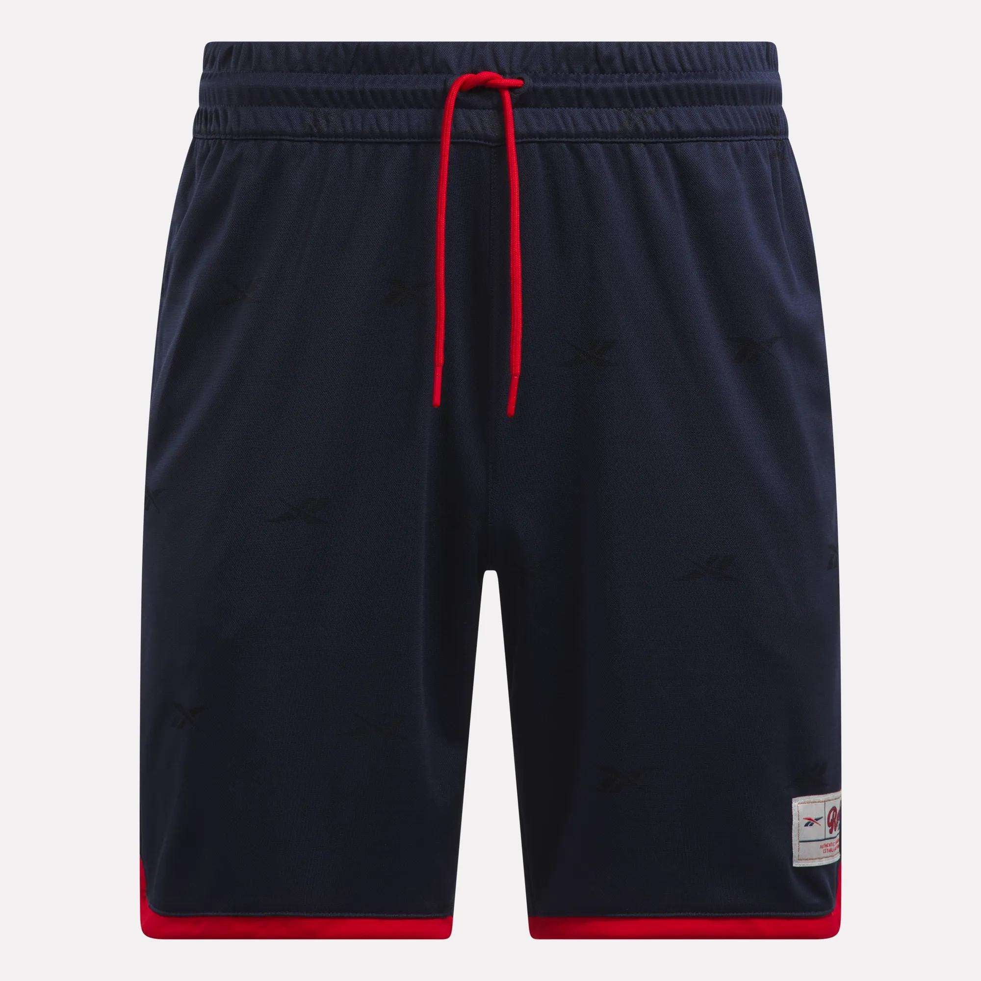The Reebok Men's United Shorts 9" are navy blue lightweight polyester shorts with red trim at the leg openings, a red drawstring, subtle logo patterns, and a small logo patch near the left hem.