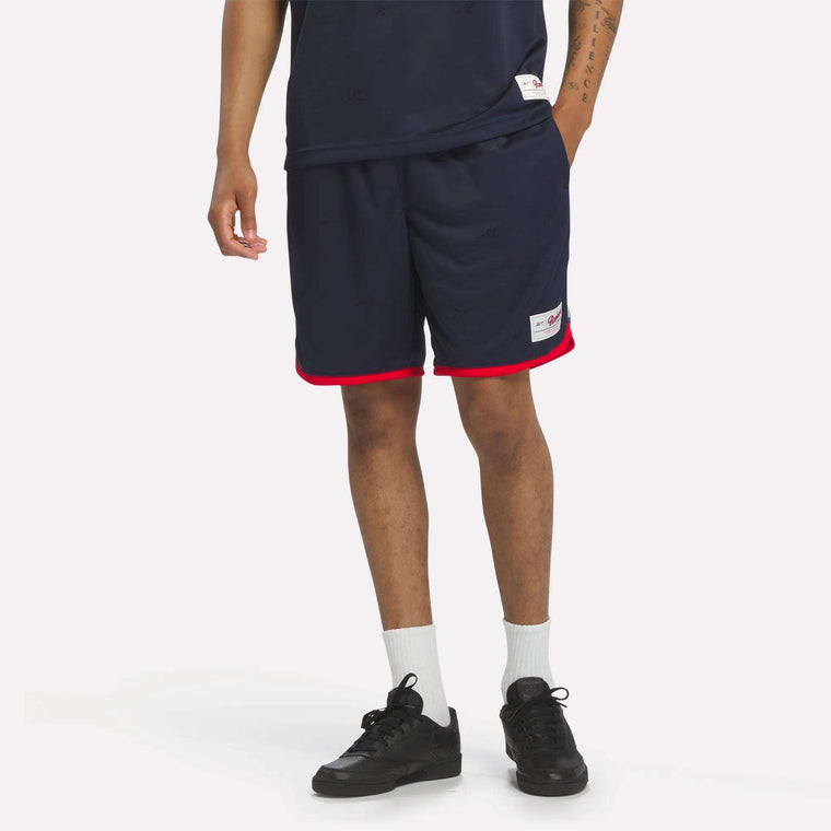 A person, shown from the waist down against a white background in sporty style, wears Reebok Men's United Shorts 9" in Vector Navy with red trim, paired with a navy shirt, white crew socks, and black sneakers.