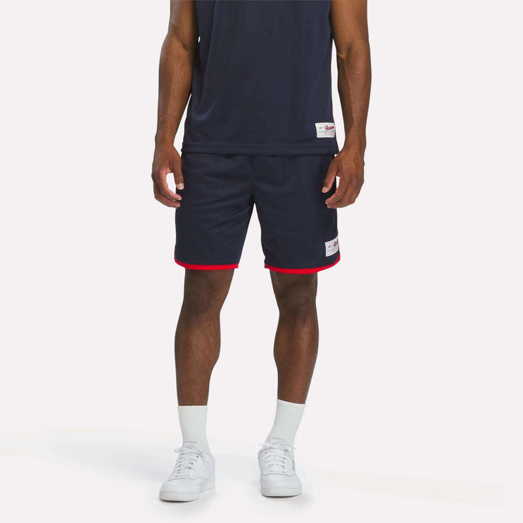 A person stands against a light background wearing Reebok Men's United Shorts 9" in navy with red trim, paired with a navy sleeveless shirt, white socks, and white sneakers. Only the lower torso and legs are visible.