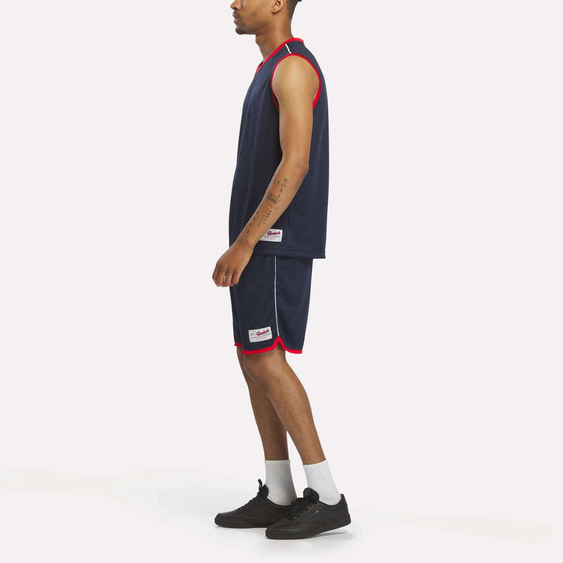 A person stands sideways in a navy blue basketball jersey and Reebok Men's United Shorts 9" with red trim, white socks, and black sneakers, showing off a sporty look against a plain white background.