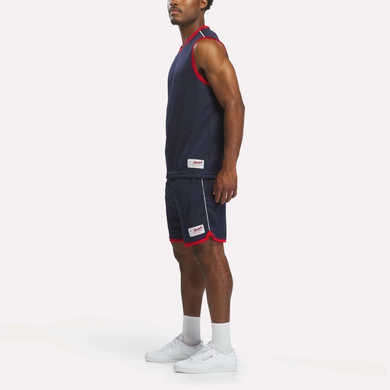 A man stands sideways wearing a sleeveless navy athletic outfit with red trim, complemented by Men's United Shorts 9" from Reebok, which are crafted from lightweight polyester for a sporty style. He pairs the look with white socks and white sneakers against a plain white background.
