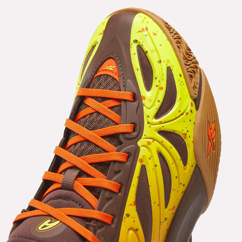 A close-up of the Reebok Reese’s x Angel Reese 1 Basketball Shoes – Big Kids highlights bright orange laces, a brown upper, yellow accents with orange speckles, mesh panels, and a rubber sole with textured tread—showcasing the dynamic style inspired by trending Angel Reese sneakers.