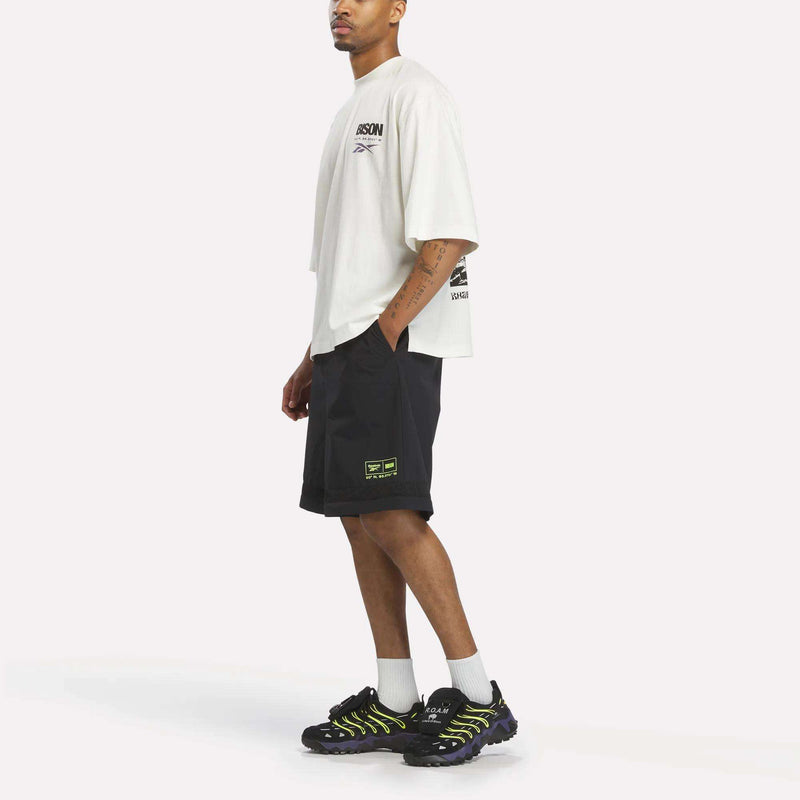 A person wears a Reebok x A Herd of Bison Transform Pants, a white t-shirt with bison graphics, durable ripstop shorts with yellow accents, and black athletic shoes with purple soles.