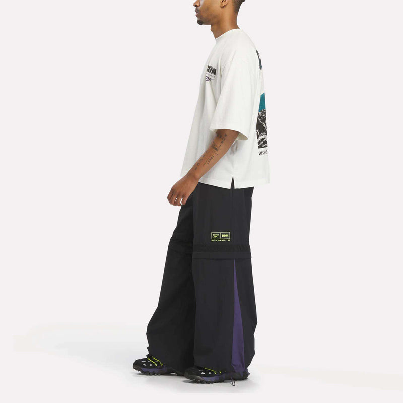 Side view of a person wearing an off-white Reebok x A Herd of Bison Graphic Tee with a bison on the back and black wide-leg trousers with purple side panels.
