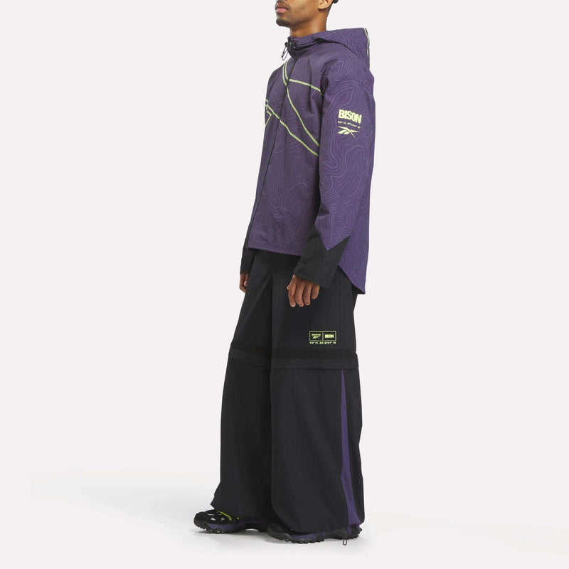 A person wears a purple Reebok x A Herd of Bison Packable Jacket with a topographical line pattern and neon green accents, featuring the word BISON and geographic coordinates, paired with loose-fitting black trousers.