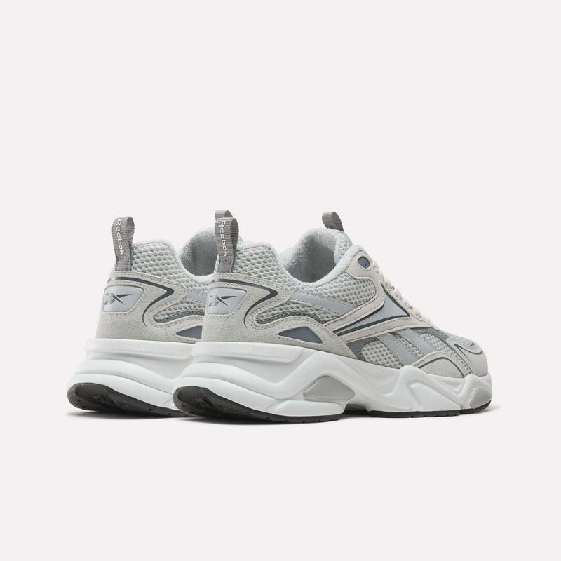 A pair of Reebok Men’s Charge Shoes in gray and white, designed with chunky soles, mesh panels, and cushioned midsoles for enhanced comfort. Featuring Reebok logos on the side and heel, these training shoes are displayed on a white background.