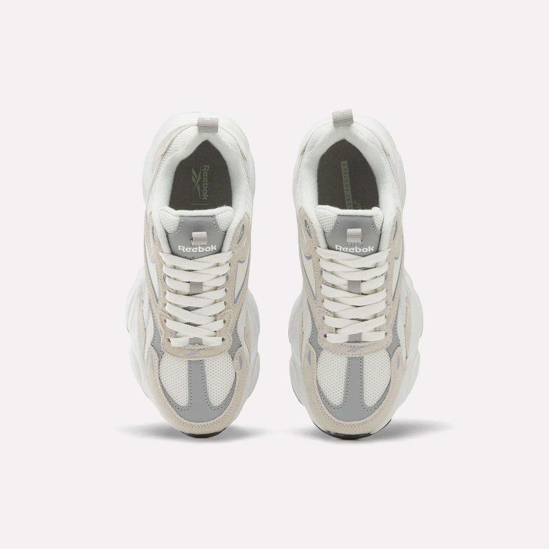 A pair of Reebok Women’s Charge Shoes in white and light gray is viewed from above, displaying the laces and the Reebok logo on each tongue against a plain white background. These women’s athletic shoes feature a subtle chunky design for enhanced style and support.