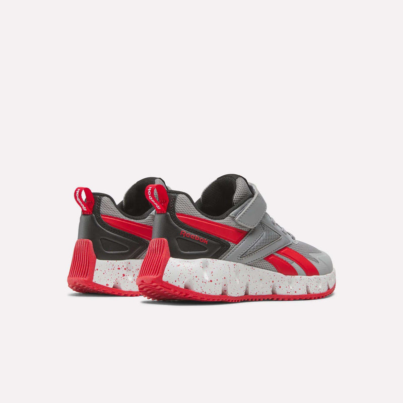 A pair of Reebok Pyro Storm 1V Shoes for little kids in gray and red, featuring white speckled soles. Shown from an angle that highlights the back and sides, these athletic shoes include red accents, Velcro straps, and pull tabs on the heels. The durable rubber outsole ensures reliable traction and long-lasting wear.