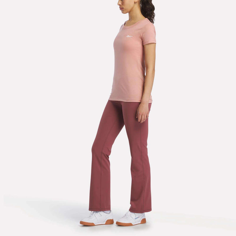 A woman stands sideways against a white background, wearing a light pink Reebok Women's T-Shirt, maroon flared pants, and white sneakers.