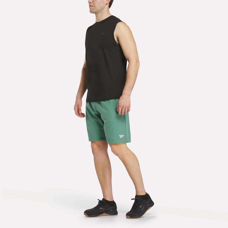A man stands against a plain white background wearing the Reebok Men's Elite Sleeveless Top, crafted from cotton flex knit, styled with green shorts and black Reebok sneakers.