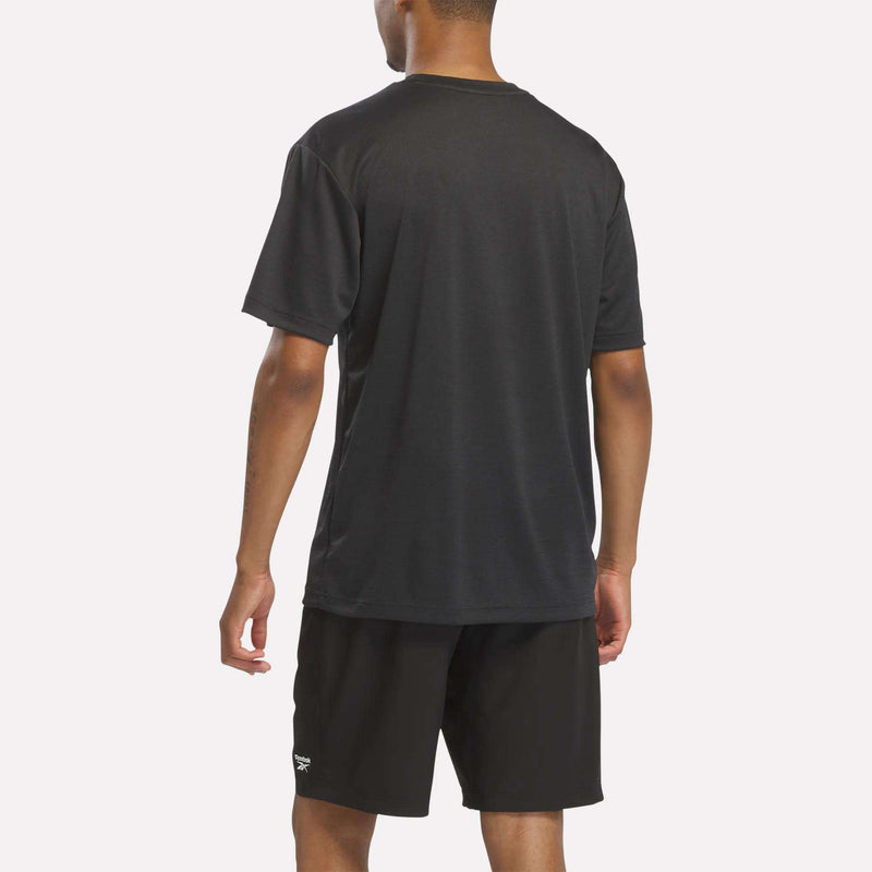 A person is standing with their back to the camera, wearing a plain black Reebok Men’s Director Short Sleeve Top and black shorts. The background is a light, solid color, emphasizing the men’s workout top and its flexible short-sleeve design.