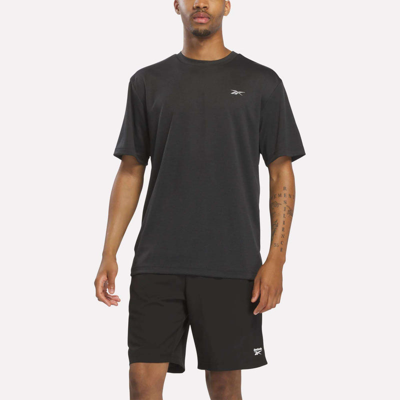 A person wearing a black heather Reebok Men’s Director Short Sleeve Top and matching black shorts stands against a plain white background. The workout top features a stretchy short-sleeve design for added comfort and flexibility.