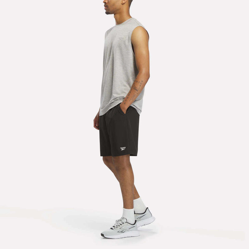 A man wearing a Reebok Men's Elite Sleeveless Top in gray, black shorts, white socks, and white sneakers stands with one hand in his pocket against a plain white background.