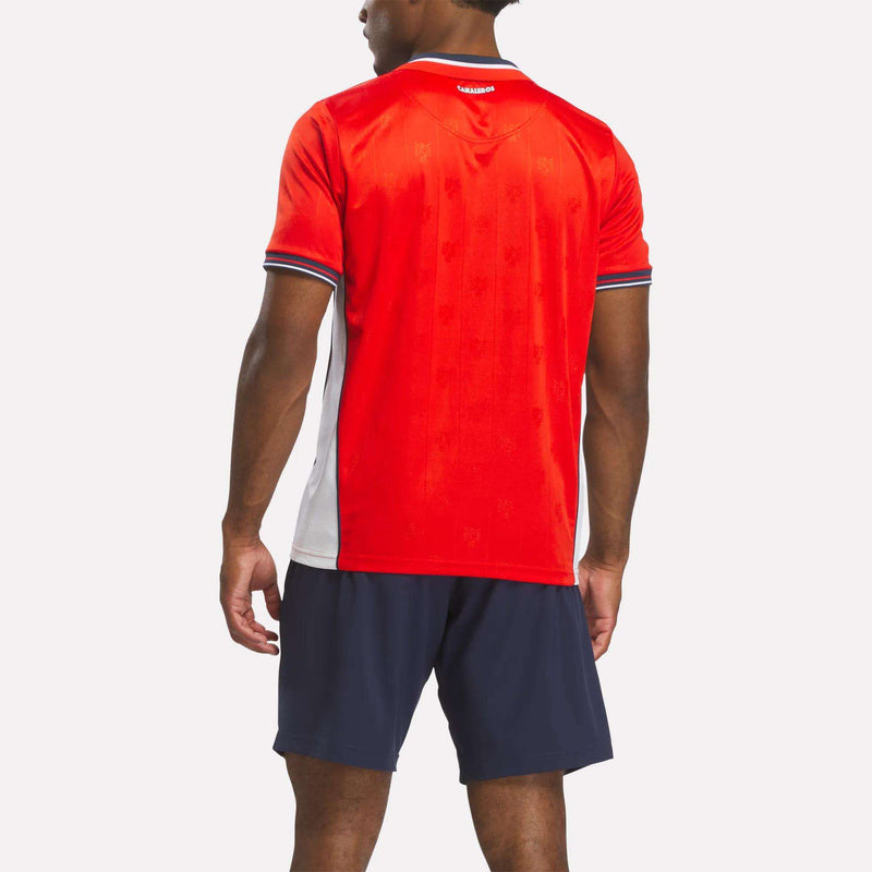 A man stands facing away, wearing the Reebok Men's Panama Home Jersey in red and white with navy shorts against a plain white background.