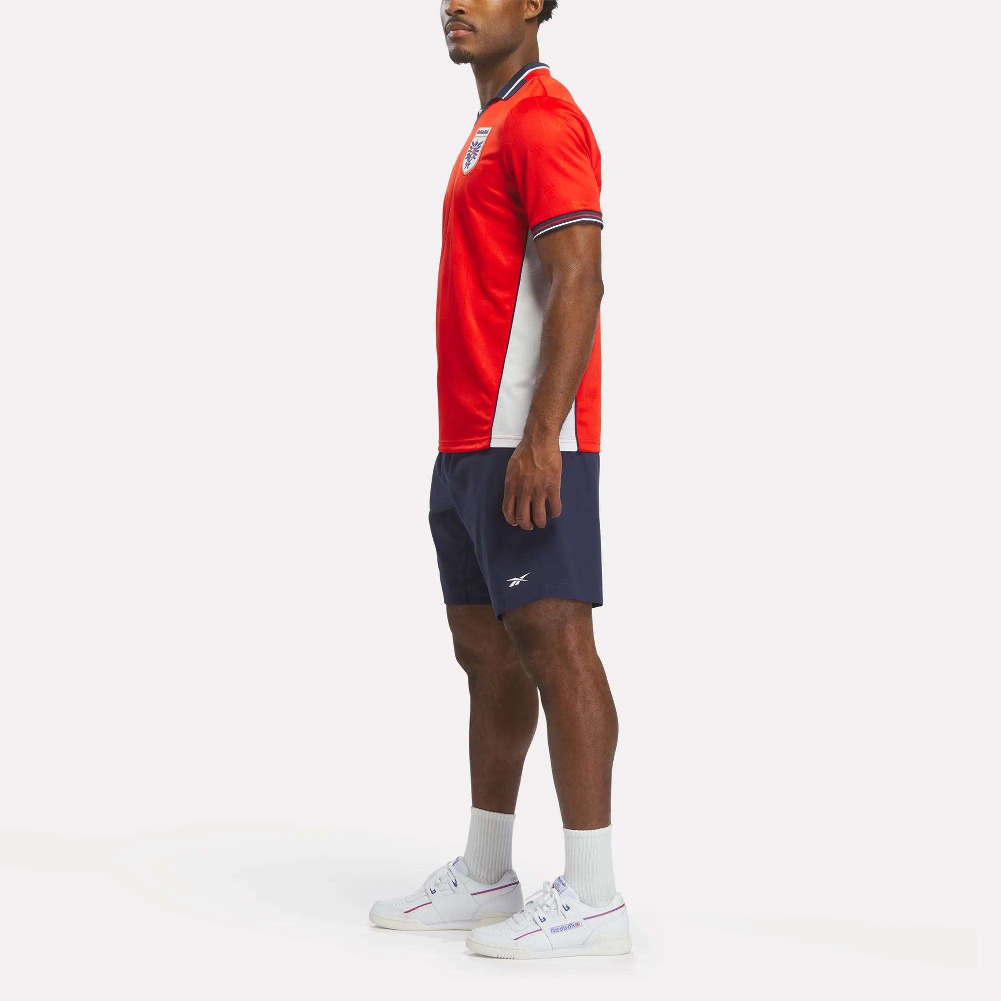 A man stands sideways in a retro-style Reebok Men's Panama Home Jersey with navy shorts, white socks, and white sneakers against a plain white background.