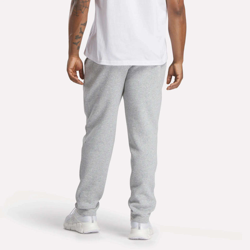 A person is shown from the waist down, wearing Reebok Men’s Patent Pants in light gray with an adjustable waistband, paired with a slim fit white T-shirt and white sneakers. One arm featuring a partial tattoo is visible. The background is plain and light.
