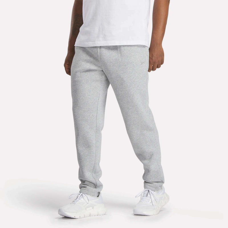 A person wearing a white t-shirt, Reebok Men’s Patent Pants in Grey Heather with a slim fit and adjustable waistband, and white athletic shoes stands against a plain background. Their face is not visible.