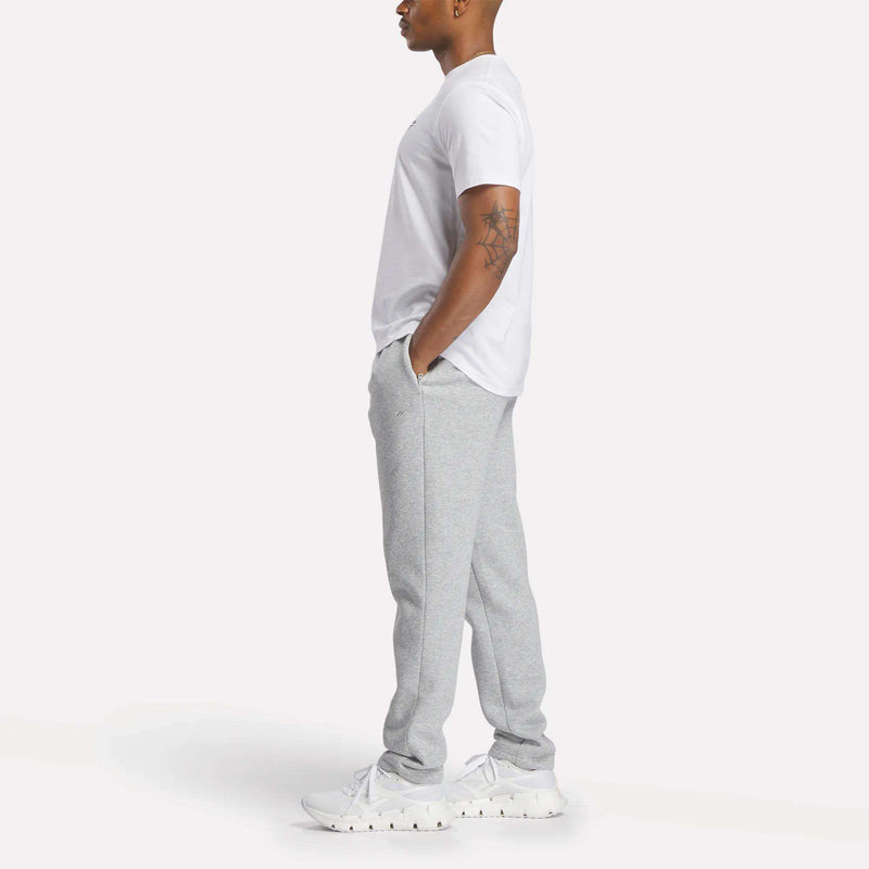 A man stands in profile, wearing a white T-shirt, the Reebok Men’s Patent Pants in a slim fit light gray design with an adjustable waistband, and white sneakers. His hands are tucked into his pockets, revealing a visible arm tattoo. The background is plain and light-colored.