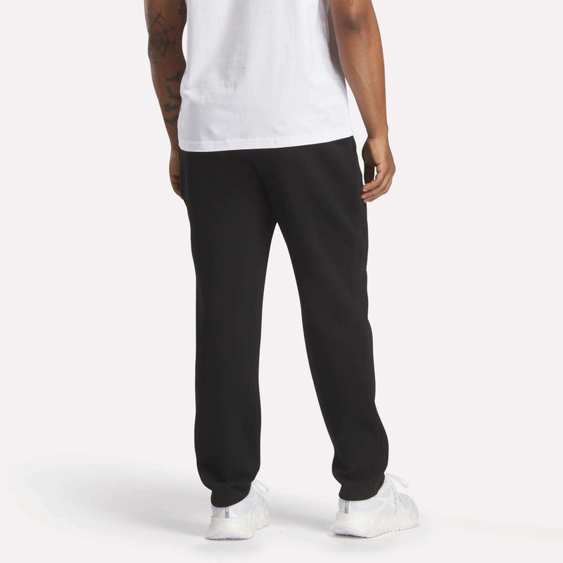 A person is standing with their back to the camera against a plain background, wearing a white shirt, white sneakers, and Reebok Men’s Patent Pants in black featuring an adjustable waistband.