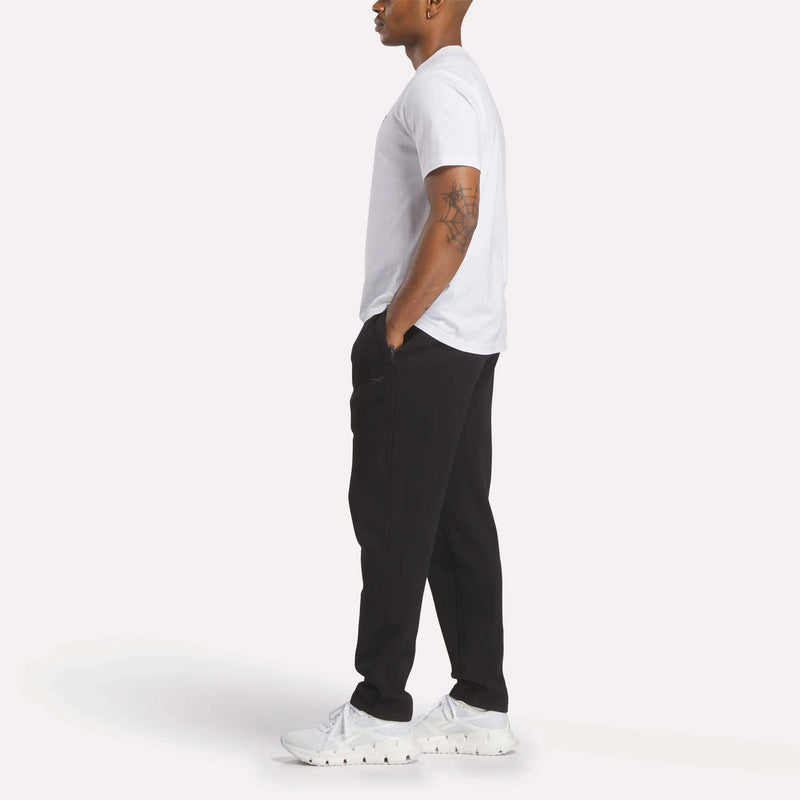 A person stands in profile against a plain light background, wearing a white T-shirt, white sneakers, and slim fit black Reebok Men’s Patent Pants featuring an adjustable waistband. Their hands are in their pockets, and a tattoo is visible on their upper arm.