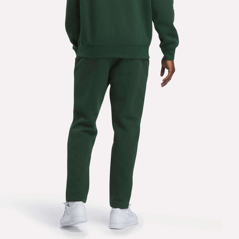 A person stands facing away, showing only the lower half of the body, wearing Reebok Men’s Patent Pants in dark green with an adjustable waistband, a matching sweatshirt, and white sneakers against a plain light background.