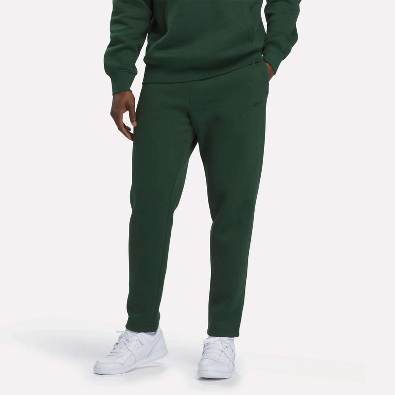 A person wearing Reebok Men’s Patent Pants in Heritage Green, styled with a matching green sweatshirt and white sneakers, stands with one hand in their pocket against a plain white background. The adjustable waistband enhances the comfort of this slim-fit, casual look.