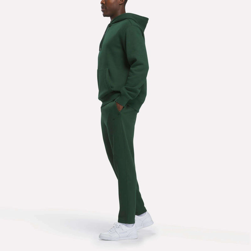 A person stands sideways against a plain white background, wearing a dark green hoodie and Reebok Men’s Patent Pants with an adjustable waistband, paired with white sneakers.