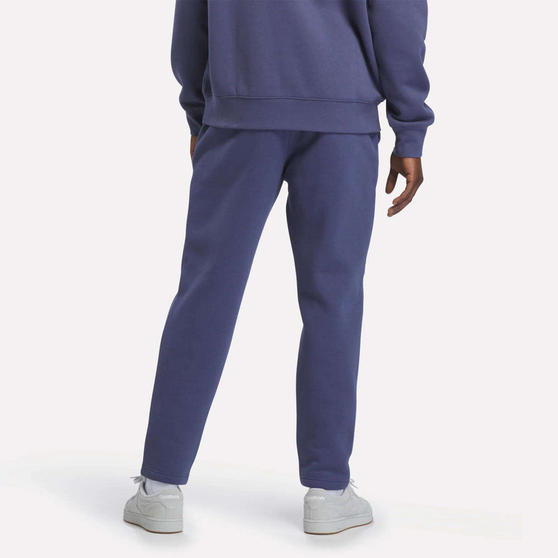 A man stands with his back to the camera, wearing Reebok’s Men’s Patent Pants—a slim fit style with an adjustable waistband—in a purple hue, matched with a coordinating sweatshirt and white sneakers. The scene features a plain light-colored background.