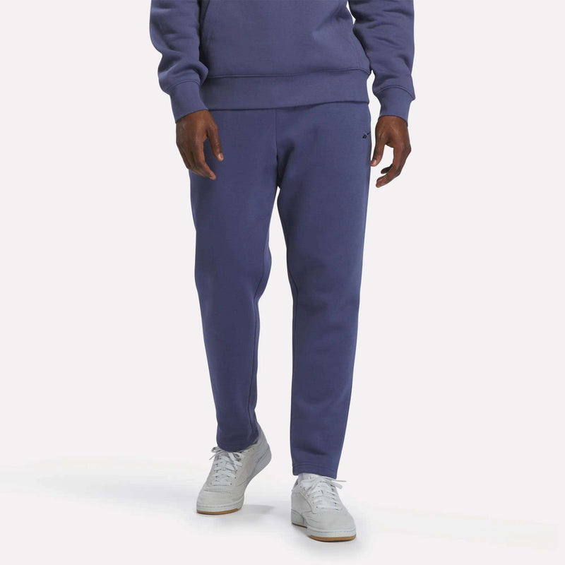 A person wearing a purple slim fit sweatshirt and Reebok Men’s Patent Pants in Legacy Blue with an adjustable waistband, paired with white sneakers, shown from shoulders down against a plain background.