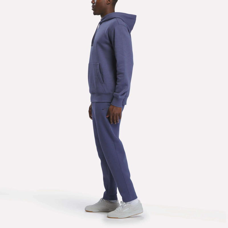 A person stands in profile, wearing a blue hoodie, Reebok Men’s Patent Pants with an adjustable waistband and slim fit, and white sneakers, against a plain white background.