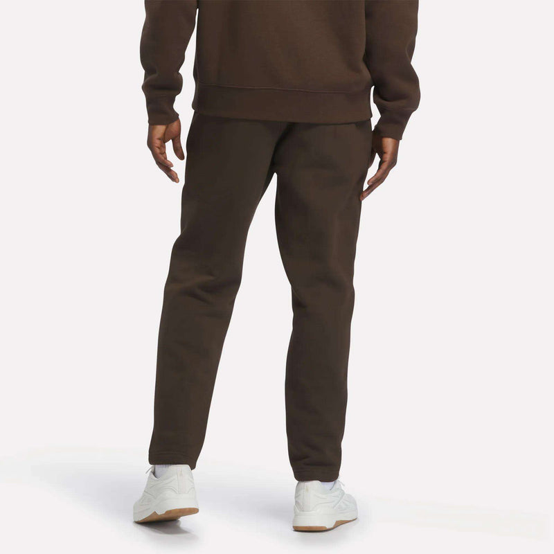 A person stands facing away, wearing Reebok Men’s Patent Pants in a brown slim fit design with an adjustable waistband, paired with a matching sweatshirt and white sneakers. The background is plain and light-colored.