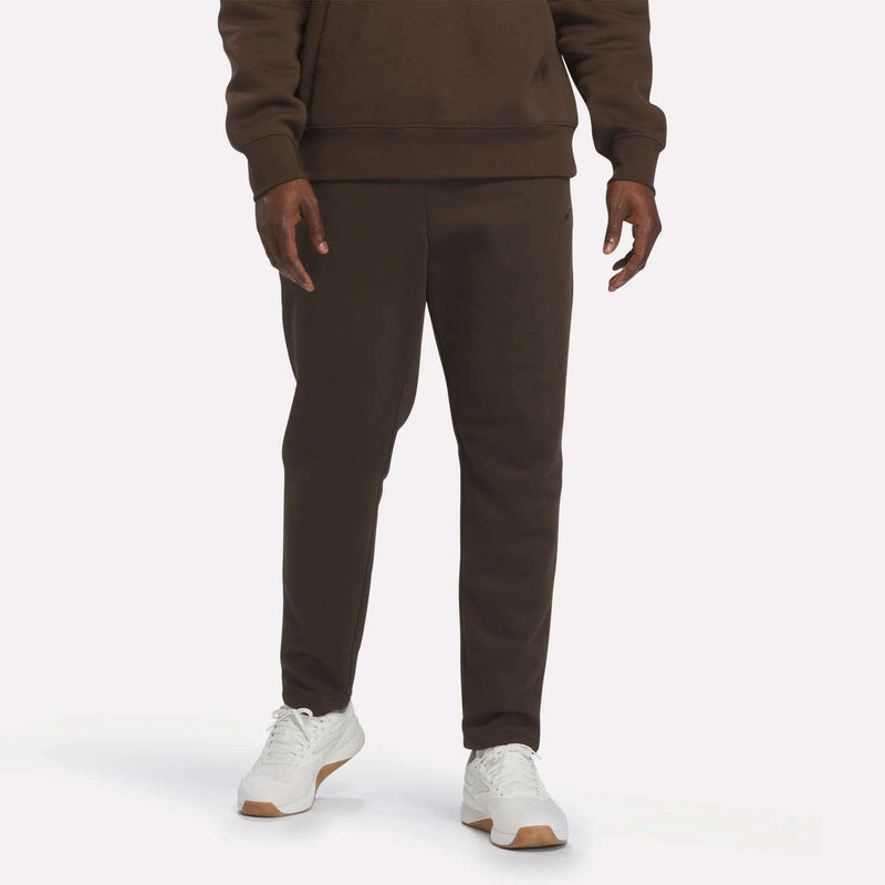 A person is shown from the waist down, wearing Reebok Men’s Patent Pants in Espresso—a slim fit style with an adjustable waistband—paired with a matching sweatshirt and white sneakers, standing against a plain, light background.