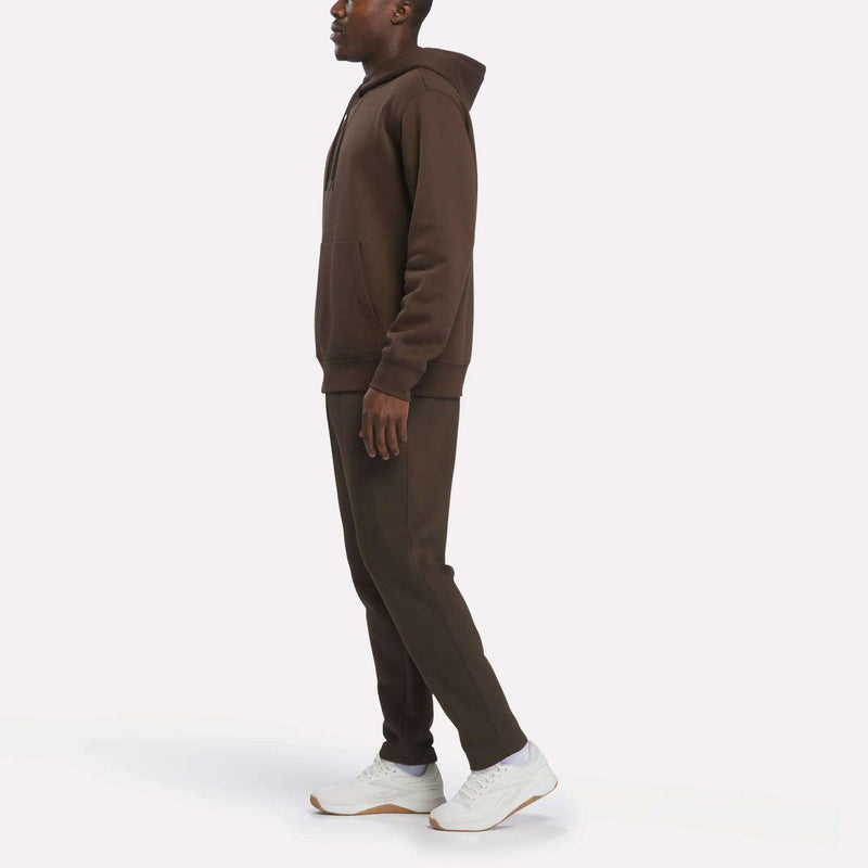 A person stands in profile against a plain white background, wearing Reebok Men’s Patent Pants—brown slim fit sweatpants with an adjustable waistband—paired with a matching brown hooded sweatshirt and white sneakers.