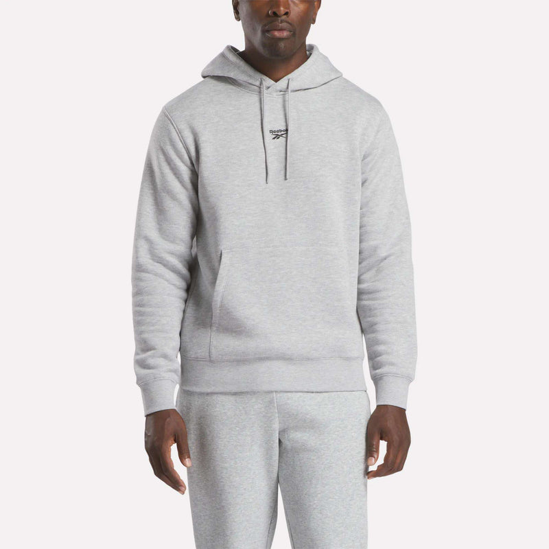 A person wearing a Grey Heather Reebok Men’s Finisher Hoodie and matching sweatpants stands facing forward. The comfortable hoodie features a front pocket and drawstrings. The background is plain and light-colored.
