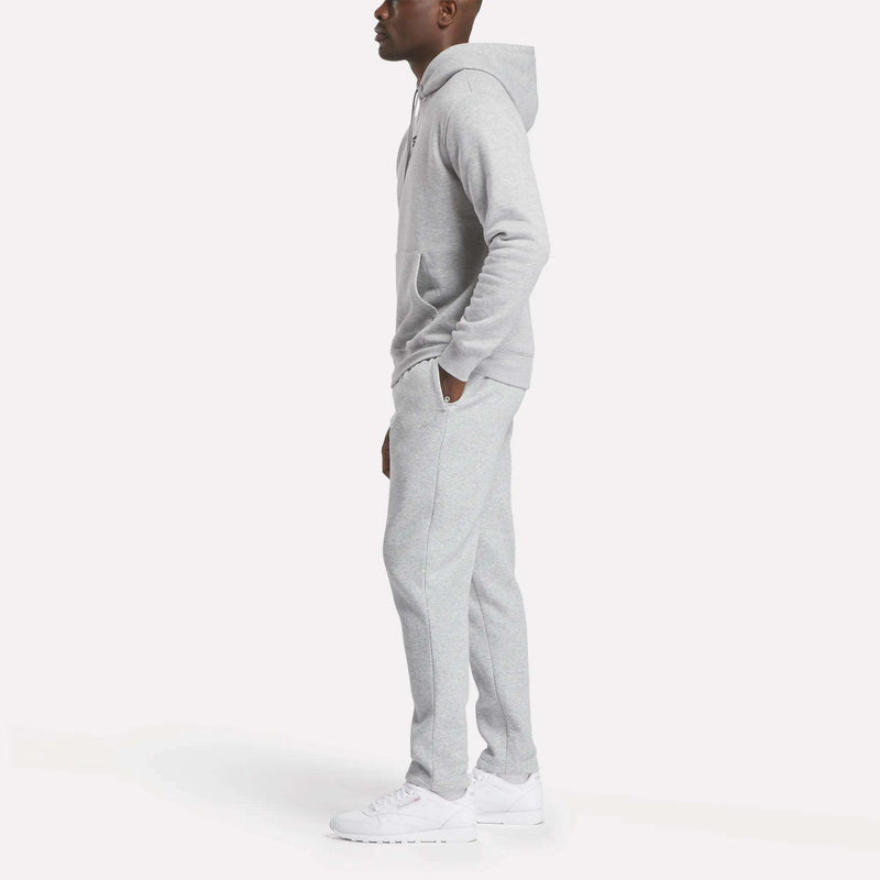 A person stands in profile, wearing the Men’s Finisher Hoodie by Reebok in light gray, paired with matching sweatpants and white sneakers against a plain white background.
