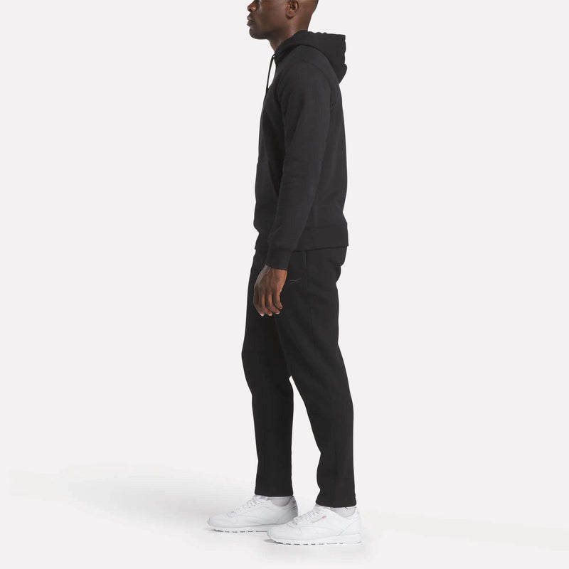 A person stands in profile, wearing the Reebok Men’s Finisher Hoodie in black, paired with black pants and white sneakers, against a plain white background.