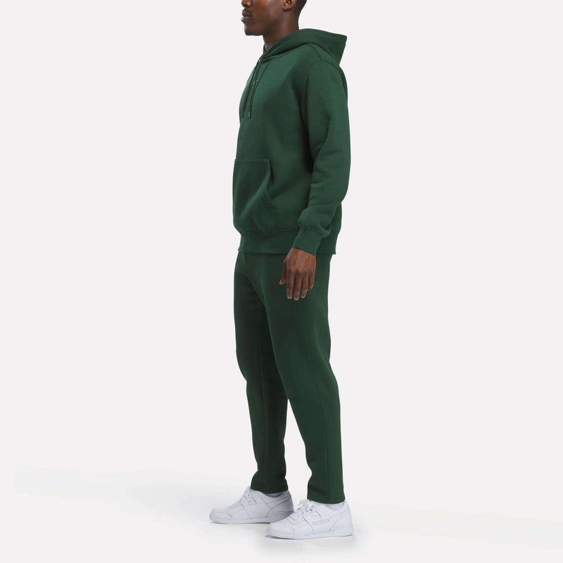 A person wearing a dark green Reebok Men’s Finisher Hoodie and matching sweatpants with white sneakers stands against a plain white background, facing left.