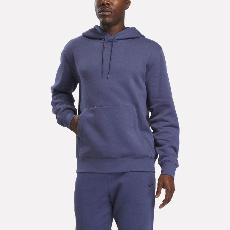 A man wears the Reebok Men’s Finisher Hoodie in purple, paired with matching sweatpants. He stands with one hand in his hoodie pocket against a plain white background.