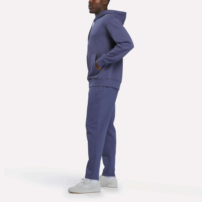 A person stands wearing the Men’s Finisher Hoodie by Reebok in blue, paired with matching sweatpants and white sneakers, looking to the side against a plain white background.