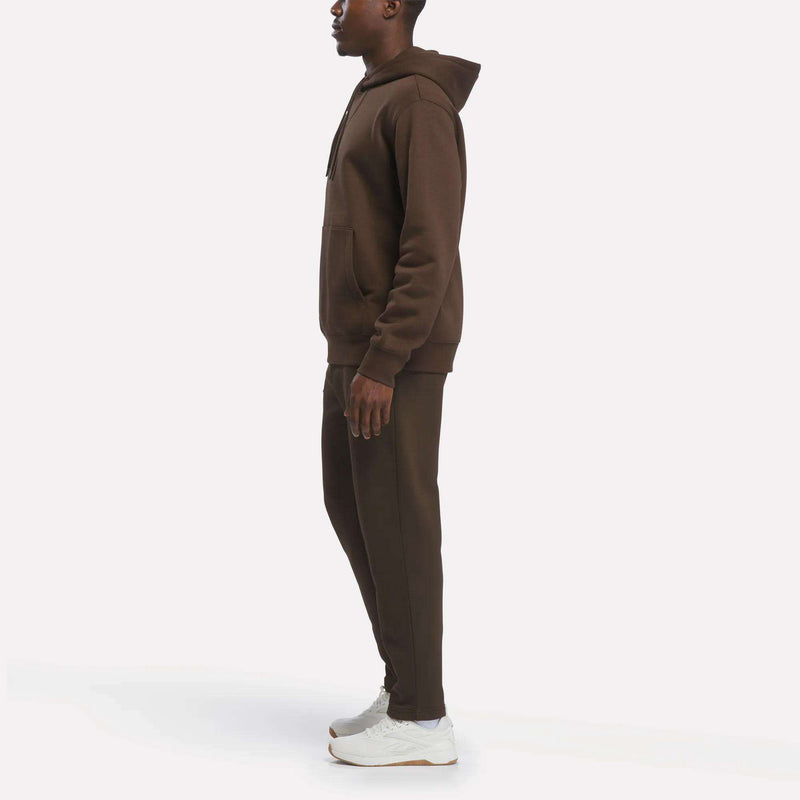 A person stands in profile wearing the Reebok Men’s Finisher Hoodie paired with matching joggers and white sneakers against a plain white background.