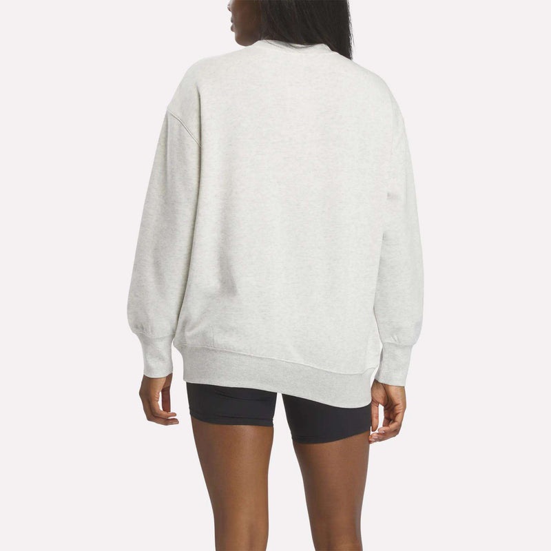 A person wearing a light gray Reebok Women’s Oversized Crewneck Sweatshirt and black shorts stands facing away from the camera against a plain white background.