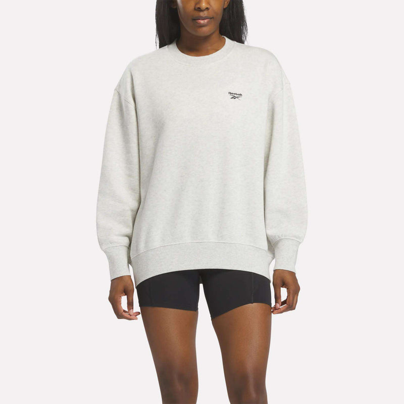A woman wearing the Reebok Women’s Oversized Crewneck Sweatshirt in Vector Chalk Heather, featuring a small logo on the chest and paired with black shorts, stands against a plain white background. The comfortable oversized design makes this sweatshirt ideal for everyday wear.