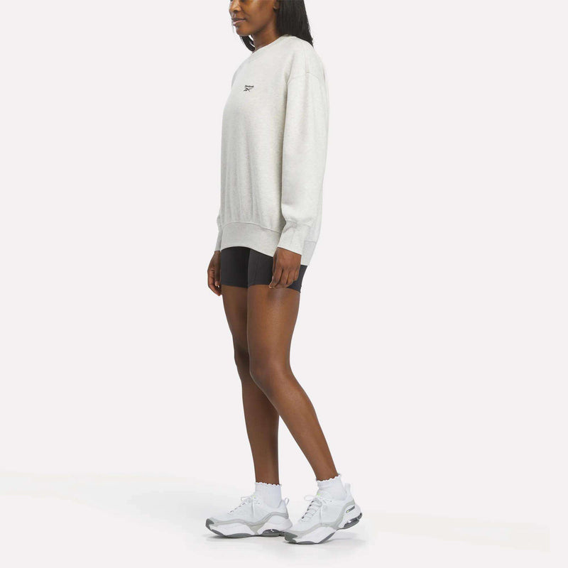A woman stands sideways against a plain white background wearing a Reebok Women’s Oversized Crewneck Sweatshirt in light gray, paired with black shorts, white socks, and white athletic sneakers. The sweatshirt features a relaxed and comfortable design ideal for casual wear.