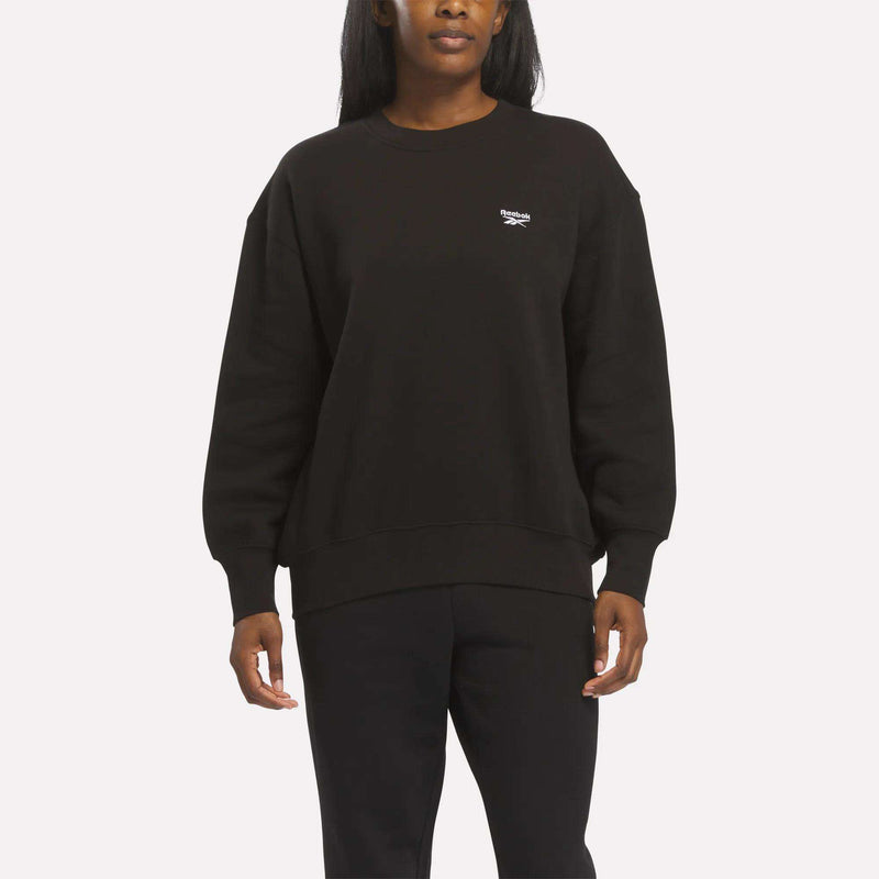 A woman is shown wearing the Reebok Women’s Oversized Crewneck Sweatshirt in black, paired with black pants, standing against a plain white background. The image displays her upper body and part of her legs.