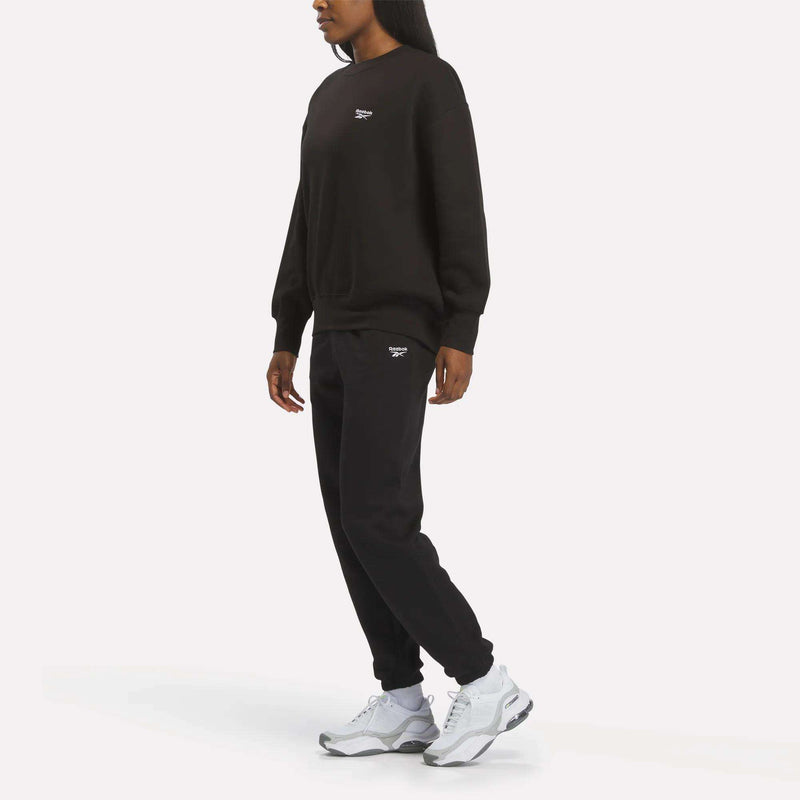 Wearing the Reebok Women’s Oversized Crewneck Sweatshirt, paired with matching black sweatpants and white sneakers, a person stands against a plain white background, highlighting the extra comfortable and relaxed silhouette of this piece.