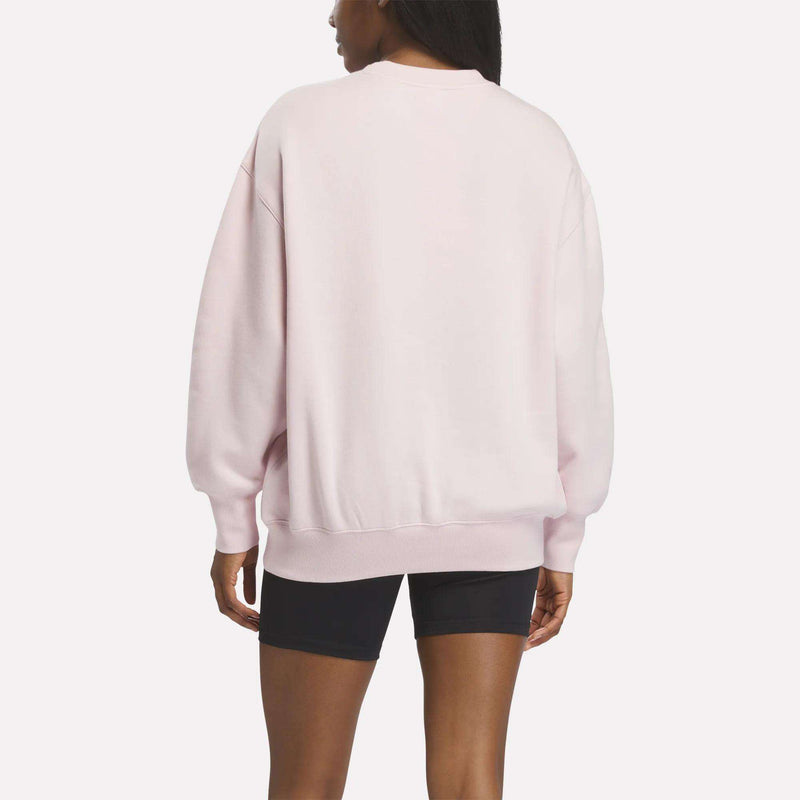 A person stands facing away from the camera against a plain white background, wearing a light pink Reebok Women’s Oversized Crewneck Sweatshirt and black shorts.
