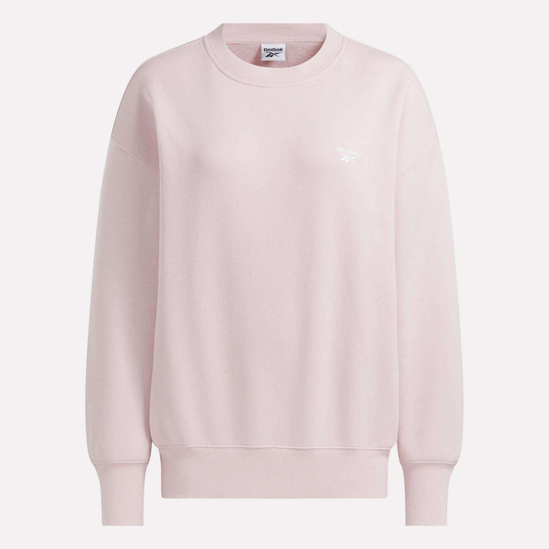 The Women’s Oversized Crewneck Sweatshirt by Reebok is shown in light pink, featuring long sleeves, ribbed cuffs and hem, and a small white Reebok logo on the left chest. This comfortable sweatshirt is displayed against a plain light background.