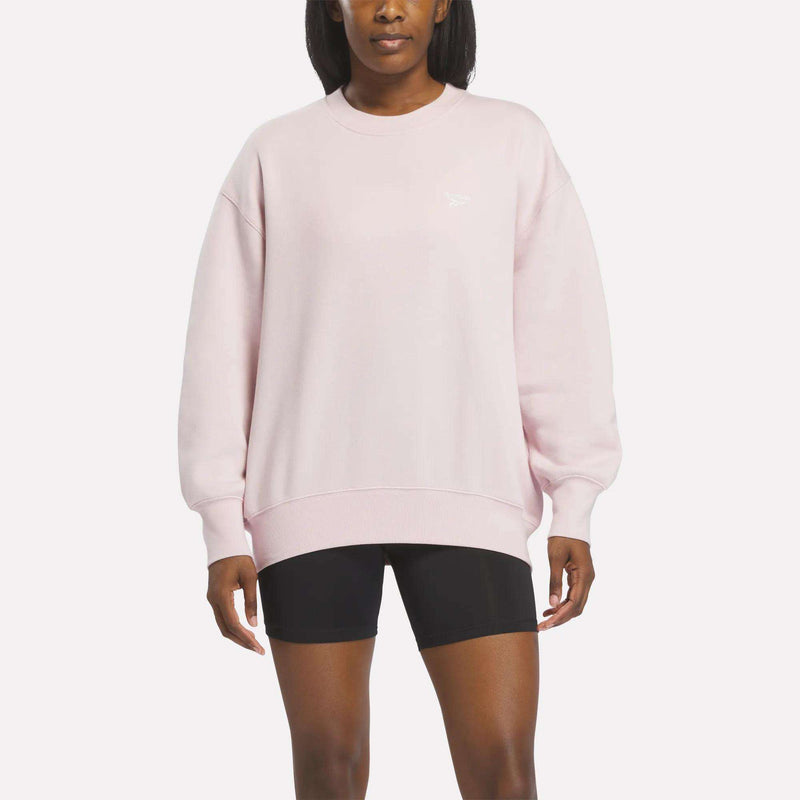 A woman stands against a plain white background, facing forward with her arms relaxed at her sides, wearing the Reebok Women’s Oversized Crewneck Sweatshirt in light pink and black shorts.