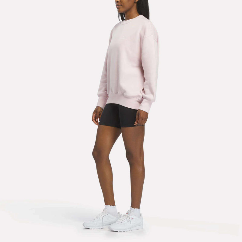 A person wearing a Reebok Women’s Oversized Crewneck Sweatshirt in light pink, paired with black shorts and white sneakers, stands against a plain white background.