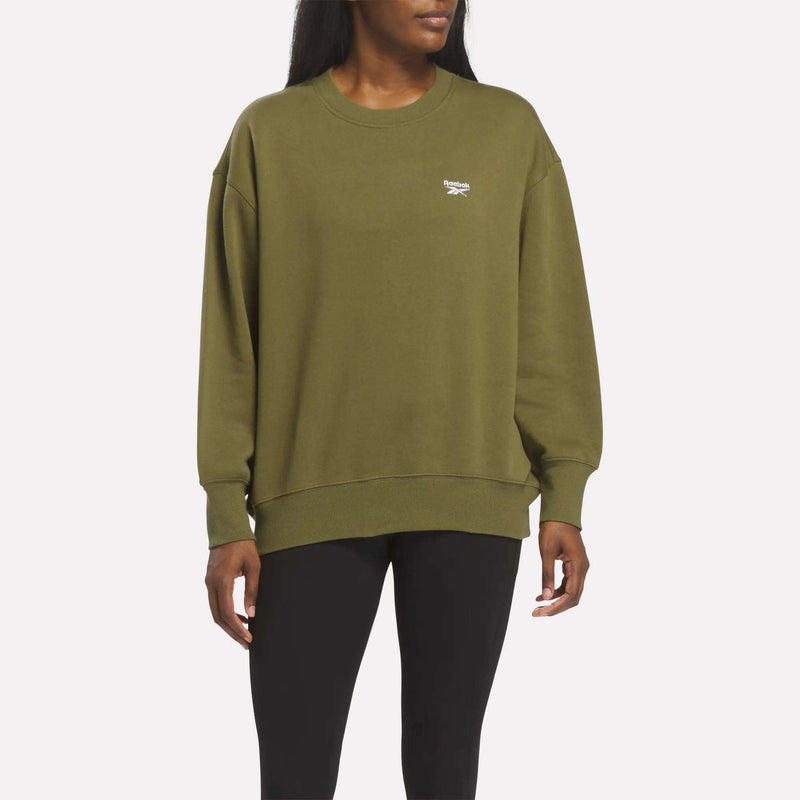 A person wears the Reebok Women’s Oversized Crewneck Sweatshirt in olive green with black leggings, standing against a plain background and facing slightly to the side.